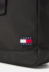 Svart stoffryggsekk med teksturert overflate, kontrasterende gummibunn og en rektangulær logo-lapp med "TOMMY JEANS" i hvitt, rødt og blått.