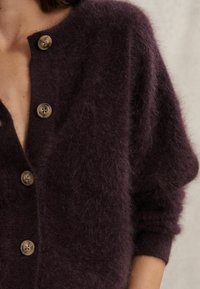 Kardigán v mocha farbe vyrobený z mäkkého a chlpatej materiálu. Má hlboký V-výstrih, oversized strih a šesť zaoblených gombíkov na prednej strane.