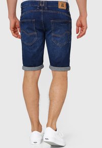 Jeansshorts mit dunkler Waschung, umgeschlagenen Säumen und zwei Gesäßtaschen mit kontrastierender Naht. Kombiniert mit weißen Turnschuhen für einen Kontrast.