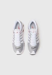 Nike Shox kedas baltā un pelēkā krāsā, ar augšdaļu no sieta un sintētiskā materiāla, melniem akcentiem, šņorēm un izteiksmīgu mīkstinātu zoli.