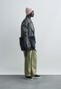 Zwarte leren totebag met een lange riem. Model draagt een grijze leren jas, lichtgroene broek met wijde pijpen en een roze gebreide muts.