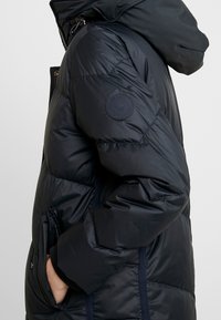 Navy puffer jacket with quilted texture, large hood, side pockets, and logo detail on sleeve. Zipper closure and ribbed cuffs.