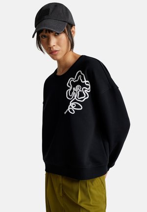 Zwarte sweatshirt met witte bloemenborduurwerk op de borst, gecombineerd met een donkere pet. Model draagt olijfgroene broek. Casual ontwerp, ontspannen pasvorm.