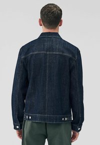 Henkilö, joka on pukeutunut tummansinisestä denim-takista, seisoo selkä katsojaan päin neutraalia taustaa vasten, mikä korostaa takin muotoilua.