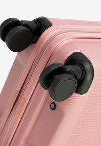 Růžový skořepinový kufr v rohu se dvěma černými otočnými kolečky, texturovaný povrch a zipový detail. Žádné další hardware nebo akcenty nejsou viditelné.