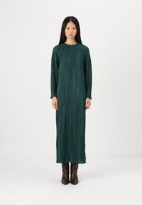 Μακριά πράσινη πλισέ φόρεμα με μακριά μανίκια και στρογγυλή λαιμόκοψη. Το ύφασμα έχει τραχεία υφή. Φορεμένο με καφέ μποτάκια με τακούνι.