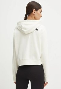 Biała krótka bluza z kapturem ze sznurkiem, z czarnym logo na górnej części pleców. Miękki materiał o gładkiej fakturze i długich rękawach.