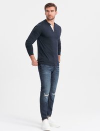 Man står med händerna bakom ryggen iförd en marinblå långärmad henleytröja, trasiga blå jeans och vita sneakers på vit bakgrund.