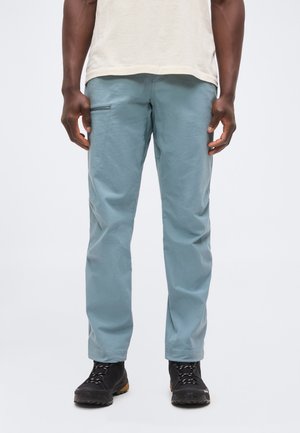 Hombre con pantalones azul claro con bolsillo lateral con cremallera, botas de senderismo negras y camisa beige sobre fondo blanco liso.