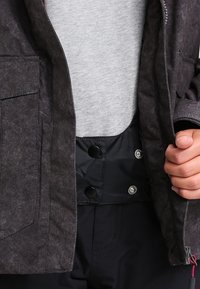 Gros plan d'une personne en train d'attacher des boutons-pression noirs sur une veste sombre portée sur une chemise gris clair.