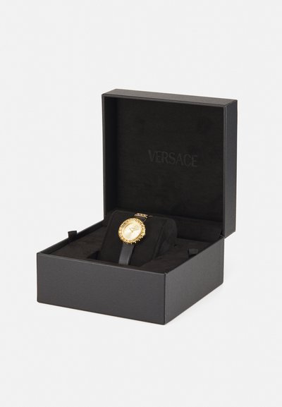 Ceas analogic din aur cu cadran rotund, curea din piele neagră și bezel texturat, expus într-o cutie neagră cu inscriptionarea "VERSACE" în interior.