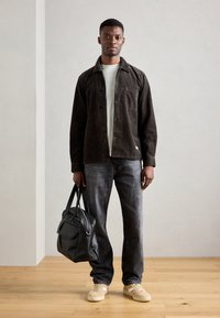 Brown corduroy shirt jacket over a light shirt, gray jeans, and beige sneakers. Holding a black leather bag. Neutral background and wooden floor.
