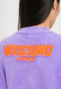 Puszysty sweter w kolorze lawendy z wyraźnym pomarańczowym napisem "MOSCHINO Jeans" na plecach. Teksturowany materiał z luźnym krojem i otwartym dekoltem.