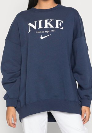 Granatowa bluza Nike z białym logo i napisem "Athletic dept. 1972", noszona przez osobę z długimi ciemnymi włosami i białymi spodniami.