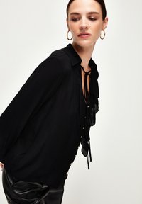 Blusa nera con colletto, parte anteriore arricciata e dettaglio con fiocco. Presenta bottoni decorativi lungo il lato e maniche lunghe e fluide realizzate in tessuto leggero.