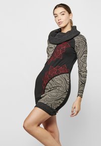Schwarzes Kleid mit langen Ärmeln, das kunstvolle goldene und rote Muster aufweist. Strukturiertes Cowl-Halsdesign und gerippten Saum an den Ärmeln und am unteren Rand.