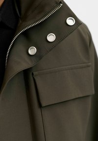 Oljčno zelena jakna z metalnim sprednjim zadrgo, pritisnimi gumbi in nalepnim žepom na prsih, izdelana iz teksturiranega blaga.