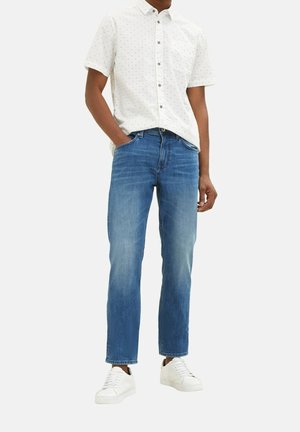 Mand iført hvid kortærmet skjorte med knapper, blå jeans og hvide sneakers, stående med den ene hånd i lommen på en ensfarvet baggrund.