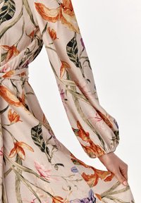 Blomstret kjole i beige stof med en tekstureret overflade, lange ærmer, bælte i taljen og livlige orange, pink og grønne mønstre.