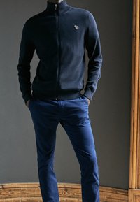 Hombre vestido con un suéter oscuro de cremallera color marino con un pequeño logo bordado y pantalones azules ajustados, de pie con las manos en los bolsillos.