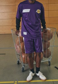 Osoba w fioletowym stroju Lakers NBA stoi obok metalowego wózka wypełnionego piłkami do koszykówki na krytym boisku.