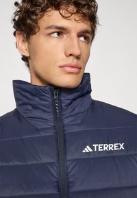 Chaqueta aislante azul marino con un cuello alto, que presenta secciones acolchadas horizontales y un logo blanco de "TERREX" en el lado izquierdo.