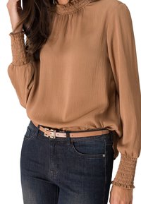 Blouse marron à manches longues avec des smocks aux poignets et à l'encolure, confectionnée dans un tissu texturé, associée à un jean bleu foncé et une ceinture claire.
