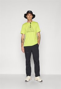 T-shirt vert vif à manches courtes avec logo Mountain Athletics noir, assorti à un pantalon sombre et un chapeau de seau noir. Baskets blanches.