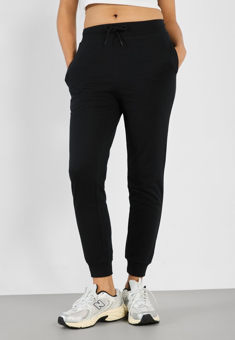 Pantaloni jogging neri con fascia elastica in vita e coulisse, caratterizzati da tasche laterali e polsini affusolati, abbinati a scarpe sportive argentate.