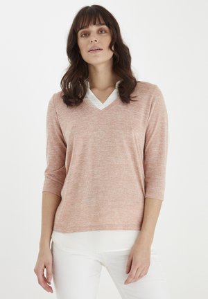 Helles rosa, strukturiertes Strickpullover mit V-Ausschnitt und drei Viertel Ärmeln, über einem weißen Hemd mit Kragen getragen. Das Model posiert frontal.