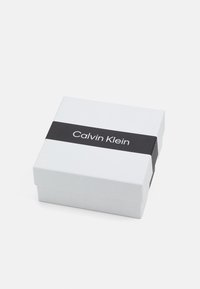Boîte en carton blanche avec une bande noire portant le texte "Calvin Klein." Texture lisse, forme carrée, design minimaliste.