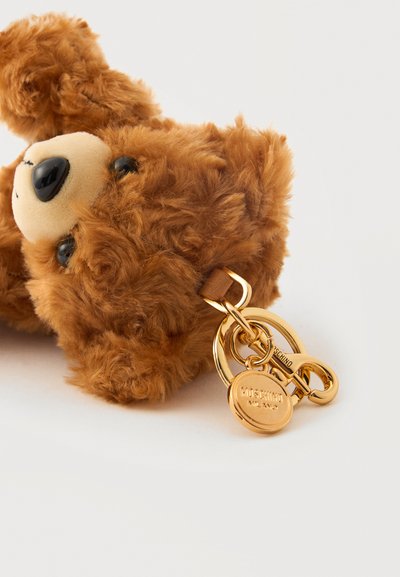 Porte-clés en peluche ours en peluche marron avec des yeux noirs, un museau beige et un anneau en métal doré avec une étiquette gravée Moschino Milano.