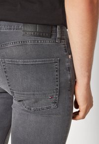 Vaqueros de mezclilla gris con un parche de cuero negro etiquetado "HILFIGER" en la parte superior de la cintura, que presentan un bolsillo trasero con un pequeño logotipo de color.