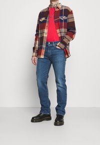 Geruite flanellen overhemd in rood, oranje en marineblauw; rood t-shirt eronder; blauwe jeans; zwarte laarzen met gele zool; casual pasvorm.