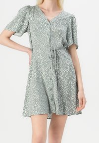 Kobieta w sukience z krótkim rękawem w kolorze szałwiowym w białe kropki, z zapięciem na guziki z przodu oraz związanym paskiem w talii, na białym tle.