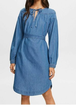 Kvinde iført en knælængde blå denimkjole med lange ærmer, en bundet udskæring og et matchende stofbælte i taljen.