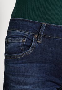 Donkerblauwe denim jeans met een getailleerde taille, voorzien van een kleine voorkant pocket, koperen klinknagels en contrasterende gouden stiksels.