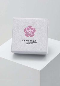 Hvid tekstureret smykkeskrin med lyserødt blomsterlogo og mærkenavnet Sensora Jewelry på en hvid piedestal.