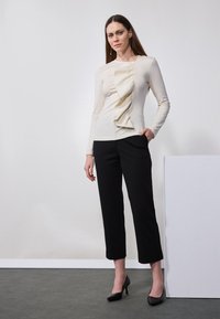 Long-sleeve cream top with a front ruffle detail, paired with black cropped pants and black high heels. Smooth texture and fitted design.