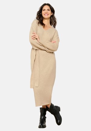 Vestido de punto beige con escote en V, mangas largas y cinturilla con lazo. Combinado con botas negras de tobillo. El vestido es de largo medio y tiene una textura suave.