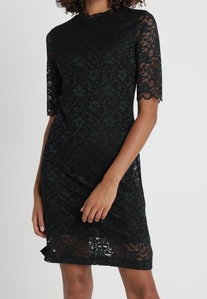 Kobieta w czarnej koronkowej sukience sięgającej do kolan, z krótkimi rękawami i wysokim kołnierzem, na jasnym, jednolitym tle.