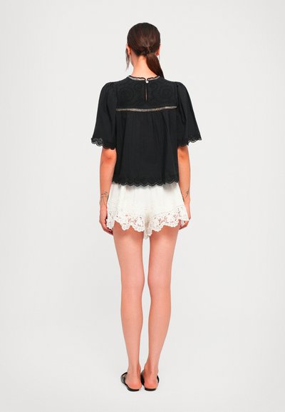 Woman standing facing away wearing a black lace-trimmed top and white lace-trimmed shorts with black flat sandals on a plain background.