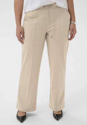 Pantalon beige à jambe droite avec des coutures frontales et une taille à cordon, porté avec des chaussures noires à bout pointu et un haut blanc.