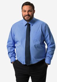 Camicia azzurra chiara con bottoni e taschino, abbinata a una cravatta scura e indossata con pantaloni neri. La camicia ha maniche lunghe e una texture liscia.