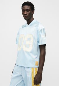 Ανοιχτό μπλε λευκό πουκάμισο από διχτυωτό ύφασμα με λαιμόκοψη σε σχήμα V, υπερμεγέθη εφαρμογή και κίτρινο αριθμό "03". Περιλαμβάνει τρεις ρίγες στα μανίκια και λογότυπο με μπαλώματα.