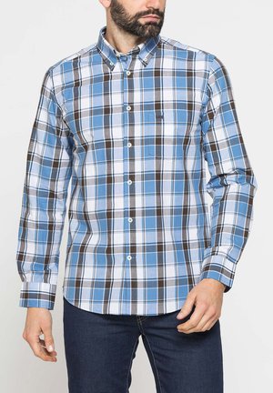 Uomo che indossa una camicia a quadri blu, bianca e marrone con bottoni e maniche lunghe, con tasca sul petto e jeans blu scuro, rivolto leggermente a destra.