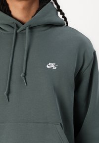 Oseba v temno zeleni Nike SB hoodieju z vrvicami in sprednjim žepom, ki prikazuje zgornji del prsnega koša in ramena.