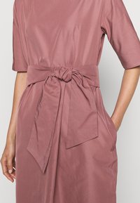 Růžové šaty s uzlovým páskem v pase, krátkými rukávy a měkkou texturou. Obsahují boční kapsy a mají uvolněný střih.