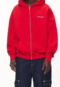 Red zip-up hoodie with a front pocket, made of cotton blend fabric. Features contrast white logo text and ribbed cuffs. Paired with dark jeans.