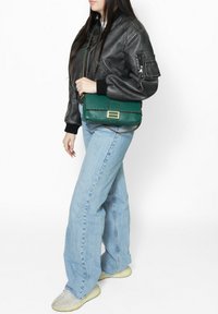 Veste bomber en cuir noir avec des poignets côtelés, jeans bleu clair à jambes larges et un sac bandoulière texturé vert avec des accessoires dorés.
